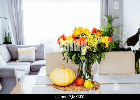 Eine Vase aus gelben und orangen Rosenblüten, frischem Kürbis, Apfel und Birne auf einer Küchentisch-Theke mit offenem Wohnzimmerhintergrund. Herbst hom Stockfoto