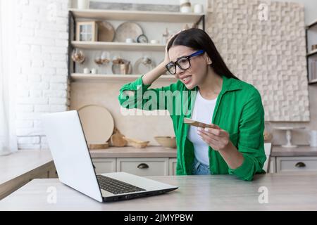 Währungskrise. Kreditschuld. Verärgert junge Frau in Brille und grünem Hemd mit Kreditkarte, sitzt am Laptop zu Hause, hält den Kopf, schockiert, verwirrt. Stockfoto