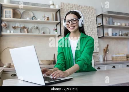 Porträt einer jungen, tausendjährigen Studentin, die zu Hause online mit einem Laptop in Kopfhörer, grüner Kleidung und Brille studiert. Er schaut lächelnd auf die Kamera. Stockfoto