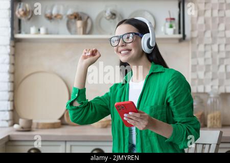 Glückliche junge schöne Frau in grüner Kleidung und Brille, die Musik in weißen Kopfhörern auf dem roten Telefon zu Hause in der Küche hört. Sie schloss die Augen und tanzte mit Stockfoto