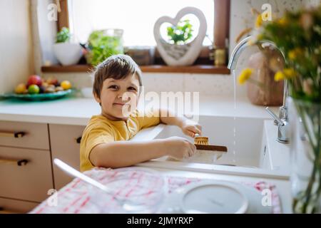 Kleiner Junge, der das Geschirr im Spülbecken in der Küche mit Holzpeeling, nachhaltige Lebenslaus, wäscht. Blick auf die Kamera und lächeln. Stockfoto