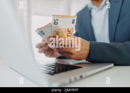 Ein Geschäftsmann mit Euro-Geld in den Händen arbeitet an einer Computertastatur am Schreibtisch, Nahaufnahme Stockfoto