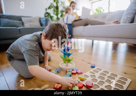 Kleiner Junge, der im Wohnzimmer mit montessori-Holzspielzeug spielt, Mutter und Schwester im Hintergrund. Stockfoto