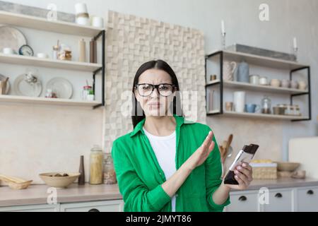 Eine junge, schöne Ernährungsberaterin mit Brille und grünem Hemd rät vom Verzehr von Schokolade ab. Er hält eine Tafel Schokolade und zeigt Nein mit der Hand. Schokolade und Süßigkeiten sind gesundheitsschädlich Stockfoto