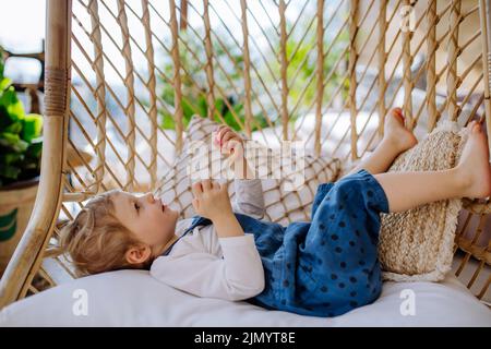 Kleines Mädchen, das zu Hause im Wintergarten liegt und spielt und auf einem Hängesessel schwingt. Stockfoto