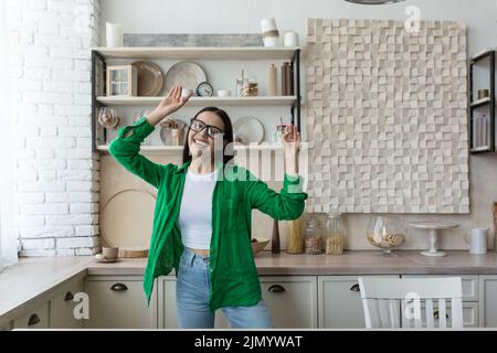 Glückliche junge schöne Frau in Brille und grünem Hemd tanzen und singen zu Hause in der Küche. Sich von den Hausaufgaben ausruhen, entspannt, die Zeit allein genießen Stockfoto