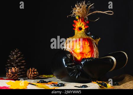 Kunstvoller Halloween-Kürbis, der auf einem schwarzen Boxhandschuh auf einigen Sackbahnen sitzt und von Spielzeugspinnen und heruntergefallenen Blättern umgeben ist Stockfoto