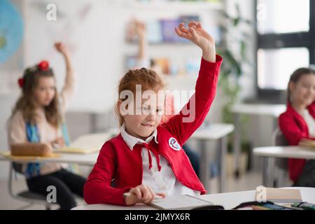 Glückliches Schulmädchen mit den Händen nach oben in der Unterrichtsstunde im Klassenzimmer in der Grundschule. Stockfoto