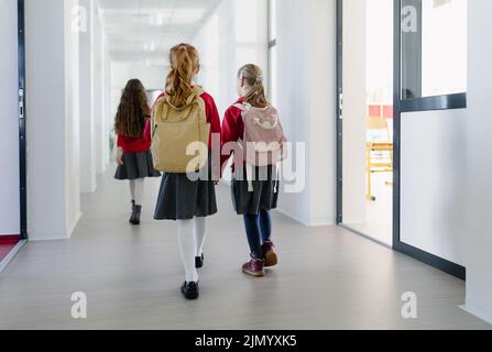 Glückliches Schulmädchen mit Down-Syndrom Klassenkameradin in Uniform, das im Schulkorridor mit Klassenkameraden läuft, Rückansicht. Stockfoto