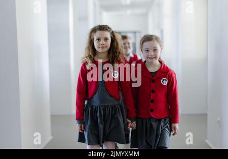 Fröhliche Schulfresserin mit Down-Syndrom in Uniform, die mit ihrem Klassenkameraden im Schulkorridor läuft und sich die Hände hält. Stockfoto