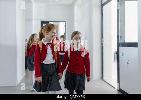 Fröhliche Schulfresserin mit Down-Syndrom in Uniform, die mit ihrem Klassenkameraden im Schulkorridor läuft und sich die Hände hält. Stockfoto