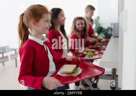 Fröhliche Schulkinder, die mit Tabletts in der Schlange stehen und in der Schulkantine zu Mittag essen. Stockfoto