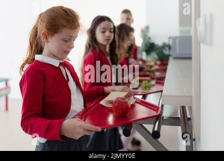 Fröhliche Schulkinder, die mit Tabletts in der Schlange stehen und in der Schulkantine zu Mittag essen. Stockfoto