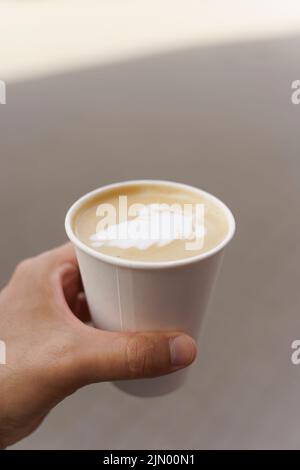 Papiertasse mit Cappuccino in der Hand auf der Straße der Stadt. Kaffeegetränk zum Mitnehmen. Hochwertige Fotos Stockfoto