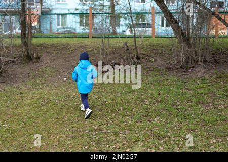 Kind läuft zum Zaun. Die Kinder haben sich auf der Straße verlaufen. Junge eilt nach Hause. Blaue Jacke. Stockfoto