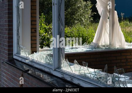 Zerbrochene Glasfenster, verursacht durch Vandalismus oder Abriss, in einer Ziegelwand eines Hauses. Die Vorhänge sind teilweise nach unten. Scharfe Spitzen des Glasstifts aus Falz Stockfoto