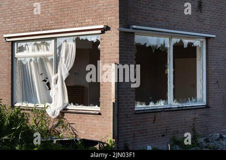 Zerbrochene Glasfenster, verursacht durch Vandalismus oder Abriss, in einer Ziegelwand eines Hauses, scharfe Punkte aus Glas Stick aus dem Falz. Vorhänge hängen heraus Stockfoto