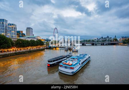 Blick Richtung Süden entlang der Themse und des Südufers in Richtung Festival Pier, London Eye und Hungerford Bridge mit festfahrenden Flussbooten in der Abenddämmerung Stockfoto