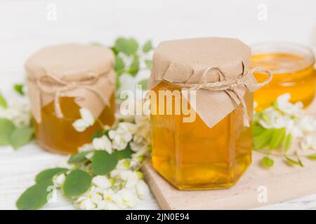 Süßes Honigglas umgab die Akazienblüten des Frühlings. Honig fließt aus einem Löffel in ein Glas. Gläser von klarem frischen Akazienhonig auf Holzgrund. Stockfoto