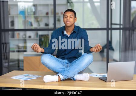 Ruhiger junger Mann, der am Arbeitsplatz im Büro sitzt und meditiert, eine Pause zu machen und stressigen Job zu vermeiden Stockfoto