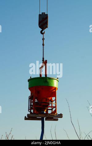 Ein farbenfroher Betoneimer hängt am Haken an Beton. Abendsonne im Winter. Haken und Ketten. Stockfoto