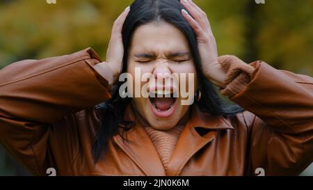 Porträt verärgerte junge hispanische Frau im Freien schreit laut in Wut wahnsinnig öffnen Mund Hände am Kopf halten Kopfschmerzen fühlt frustrierte Schmerzen Stockfoto