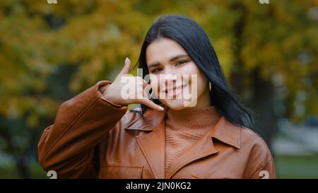 Nahaufnahme junge hübsche hispanische Frau, die im Park im Freien posiert und auf die Kamera schaut, die Handbewegung zeigt Telefon in der Nähe des Ohrs, das Zeichen zeigt, rufen Sie mich an Stockfoto