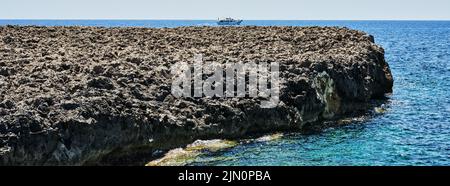 Wunderschöne felsige Küste der Insel Menorca mit türkisfarbenem mediterranem Wasser und einem fernen Boot unter klarem blauen Himmel, Spanien. Stockfoto