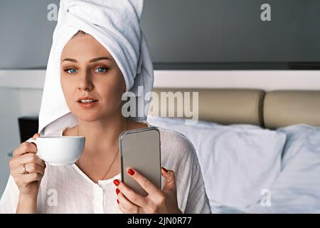 Porträt einer jungen Frau, die die Kamera anschaut. Beim Lesen von Nachrichten per Telefon, Scrollen in sozialen Medien, die mit einem weißen Handtuch auf dem Kopf im Schlafzimmer auf dem Bett sitzen Stockfoto