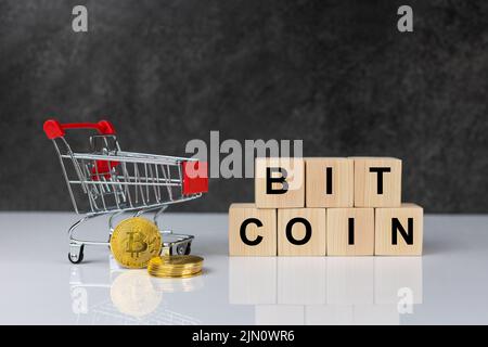 Bitcoin im Einkaufswagen und Holzblöcke mit dem Wort Bitcoin auf dunkelgrauem Hintergrund in weißem Tisch. Konzept der zukünftigen Währung. Stockfoto