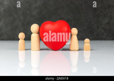 Vier kleine Holzfiguren, die eine Familie symbolisieren - eine Mutter, ein Vater und zwei Kinder, in der Nähe eines großen roten Herzens. Das Konzept der Liebe und Fürsorge der Familie. Stockfoto