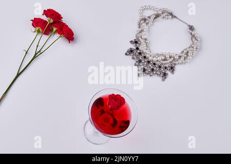 Flache Damen Accessoires auf weißem Hintergrund. Roter Cocktail mit Rosenblüten und Halskette. Stockfoto