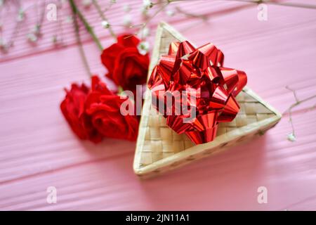 Draufsicht Triangle box mit roter Schleife. Rote Rosenblumen auf rosafarbenem Holzschreibtisch. Stockfoto