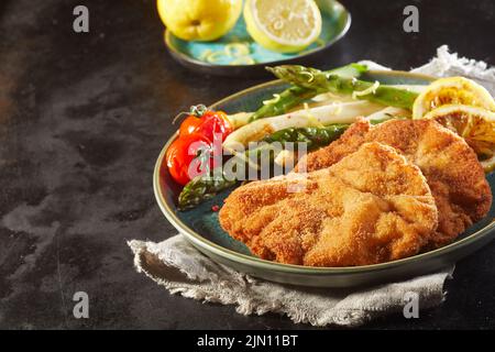 Hkeramische Platte aus Schnitzel und Spargel mit Kirschtomaten und Zitronen auf Leinenserviette auf schwarzem Hintergrund gelegt Stockfoto