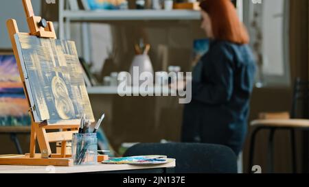 Abstrakte Malerei schwarze Leinwand mit goldenen Zeichnungen auf Staffelei steht auf dem Tisch in Werkstatt-Studio mit Pinsel rothaarige unkenntlich Frau Stockfoto