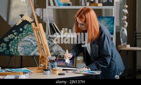 Inspiriert beschäftigt konzentriert Rotschopf Mädchen Künstlerin Frau Maler im Kunststudio Hintergrundfarben mit breiten Pinsel auf Leinwand mit Acryl-Öl bunte Farbe legt Stockfoto