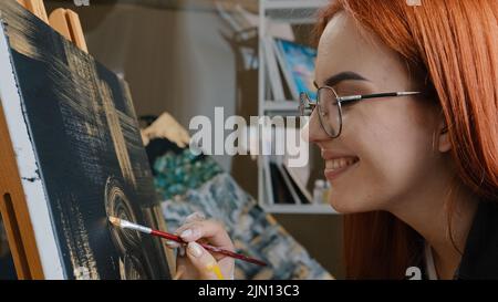 Seitenansicht Nahaufnahme rothaarige 20s Frau talentierte Künstlerin Mädchen in Gläsern weibliche Malerin malt Bild auf schwarzem Hintergrund mit goldenem Öl Acryl Dame Stockfoto