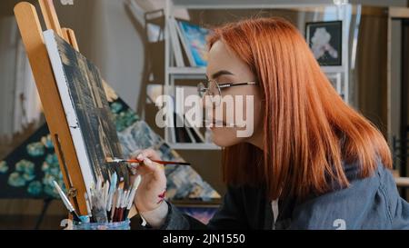 Inspiriert lustig fokussierte Künstlerin Frau mit roten Haaren Mädchen Maler trägt Gläser hält Malerei Pinsel in Mund Farben mit Ölfarben auf Leinwand auf Staffelei Stockfoto