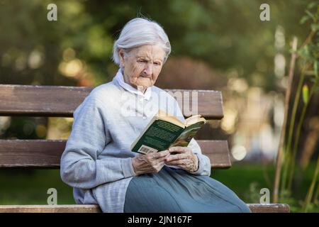 Porträt einer älteren Frau auf der Bank im Park, die ein Buch liest Stockfoto
