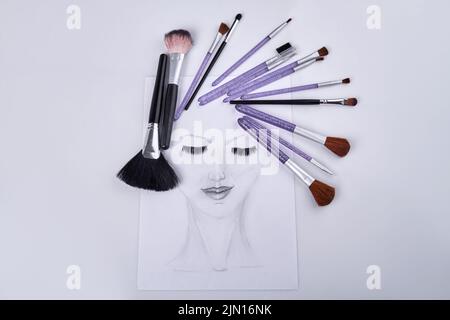 Handgezeichnetes weibliches Gesicht mit geschlossenen Augen auf weißer Oberfläche. Set aus verschiedenen lila Make-up Bürsten. Stockfoto