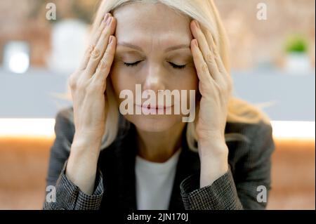 Nahaufnahme einer erschöpften kaukasischen Geschäftsfrau mittleren Alters, die Müdigkeit von der Arbeit, Überlastung, Kopfschmerzen, Migräne, Massagen in ihren Schläfen, Schließt ihre Augen, braucht Ruhe Stockfoto