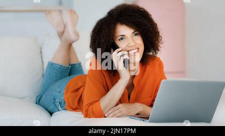 Lächelndes afroamerikanisches Mädchen mit lockigen Haaren, die zu Hause liegen und sich auf dem Telefon ausruhen und auf den Laptop-Bildschirm schauen, um sich zu entspannen, als freiberufliche Frau zu machen Stockfoto
