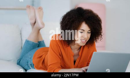 African 20s Lady 30s Frau freiberuflich mit lockigen Haaren auf dem Sofa liegend entspannendes Tippen auf Laptop macht Online-Kauf im Internet-Shop App arbeiten Stockfoto