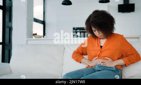 Gestresste afroamerikanische Frau auf dem Sofa sitzend berühren Bauch krank Biracial lockiges Mädchen an Magenschmerzen leiden. Unglückliche Dame, die Schmerzen empfinden kann Stockfoto