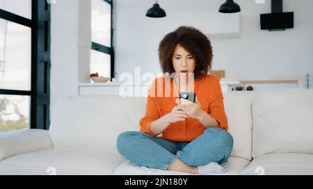 Überglücklich afroamerikanische Frau erhalten gute Angebot Gelegenheit Eingabe Smartphone Gewinn Geldpreis in mobile App sitzen auf Couch zu Hause. Glückliche Frau Stockfoto