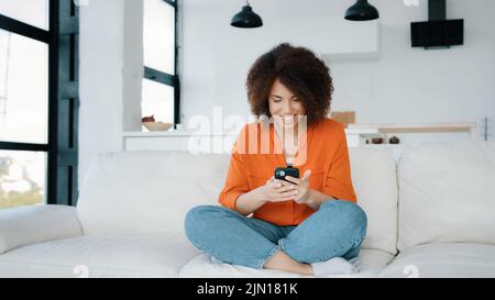 Afroamerikanische Frau sitzt zu Hause Couch suchen Smartphone mit Handy spielen mobile Spiel Sieg Reaktion erhalten gute Angebot Gelegenheit Nachrichten Stockfoto