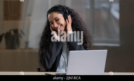 Glückliche junge arabische Frau Freiberuflerin in Kopfhörern lautes Musikhören in Kopfhörern, die im Büro frei singen und sanfte Handbewegungen genießen Stockfoto