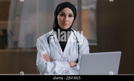 Glücklich lächelnd arabische Krankenschwester islamischen Arzt Therapeut tragen weißen Uniform Mantel Hijab sitzt in der Klinik muslimische weibliche Praktikantin Praktikantin gekreuzt posieren Stockfoto