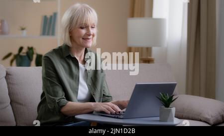 Alte Frau sitzt auf der Couch im Heimbüro beschäftigt bei der freiberuflichen Arbeit mit Laptop ältere Dame schreiben E-Mail Text in Computer-Web-Anwendung Stockfoto