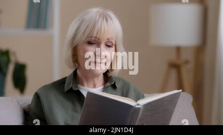 Inspirierte verträumte kaukasische alte Frau ältere Dame ältere Großmutter sitzt auf dem Sofa entspannen zu Hause Lesen Buch Roman Literatur lernen älter Stockfoto